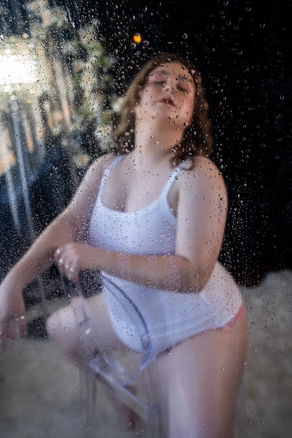 Jesse sits backward in a clear plastic chair, her head tilted back. She is seen through water droplets and haze.. She wears a wet white tank top which is transluscent and pink lacey panties. Her hands reach back and rest on her ass. Credit: Luna Sanchez, Boudoir by Hannah Rose. 